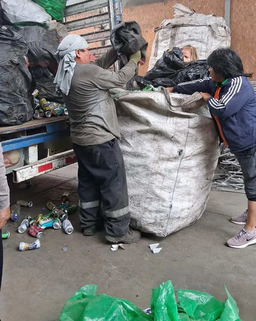 Dos recicladores cargando una bolsa de material en un camión, con latas de aluminio en el suelo